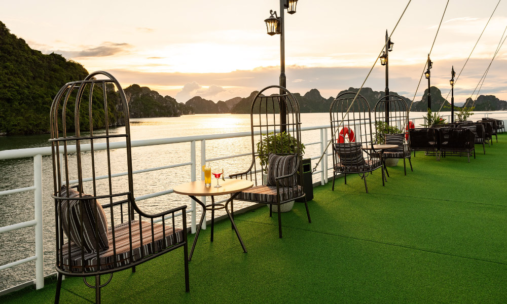 One side of Carnival sundeck with grass floor and lounge chairs on the sides, facing Halong Bay islands during sunset.