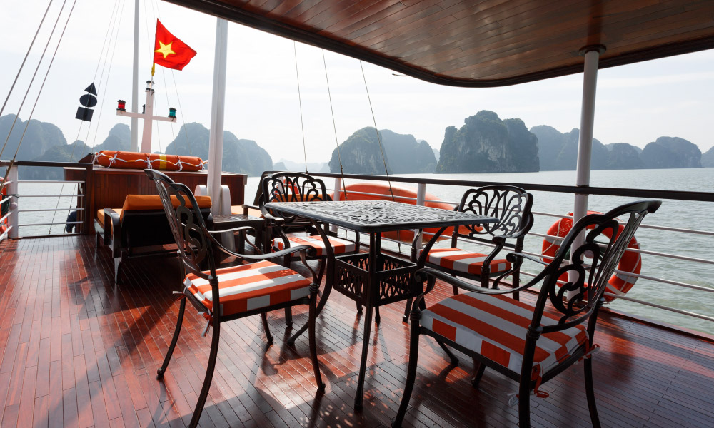 A table with four chairs on the deck of Emeraude Premium Cruise with Halong islands behind.