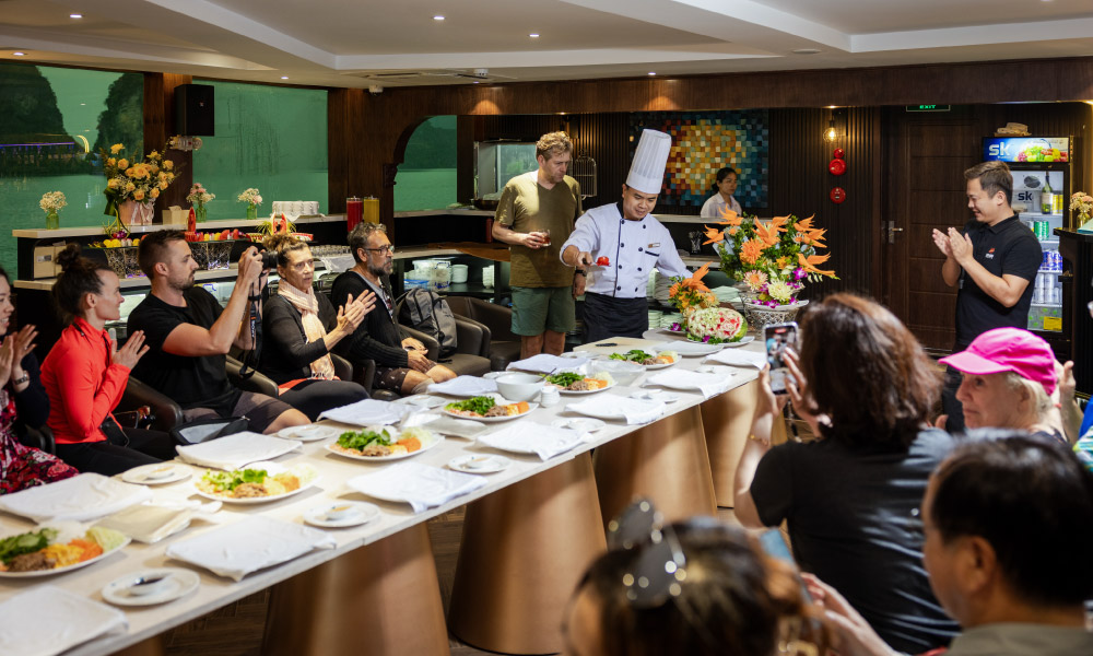 Guests hearing a chef surrounding a table with ingredients on it.