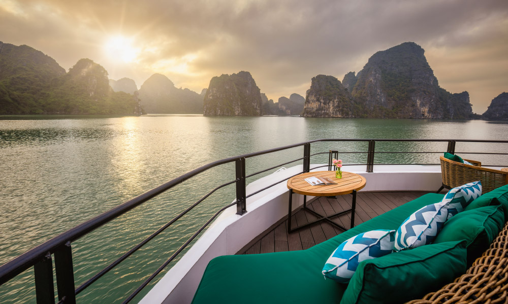 Halong Bay sunset looking from a corner deck with green sofas and a small coffee table.