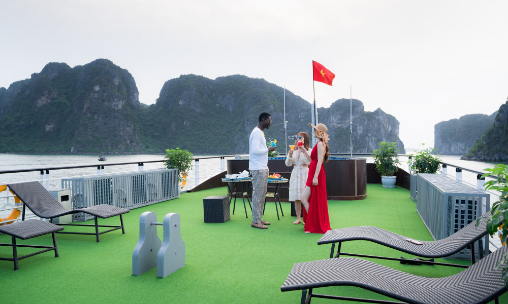 Three people standing and talking on a green sundeck, with view of Halong Bay islands surrounding the cruise.