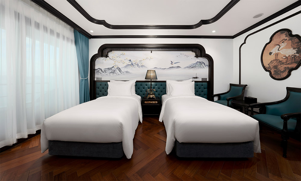 A room with two twin beds parallel, with a window on the left with the drapes down, and two chairs on the right.