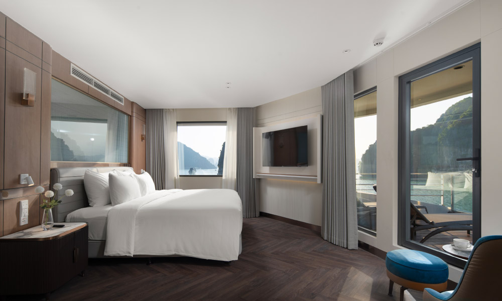 Overview of a grey room with a bed facing the TV, a bedside table with white flowers, and doors leading to a balcony outside.