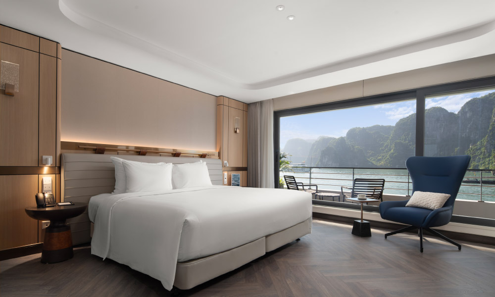 A room with double bed on the left and a blue chair at the foot, outside the windows is a balcony with view of Halong islands.