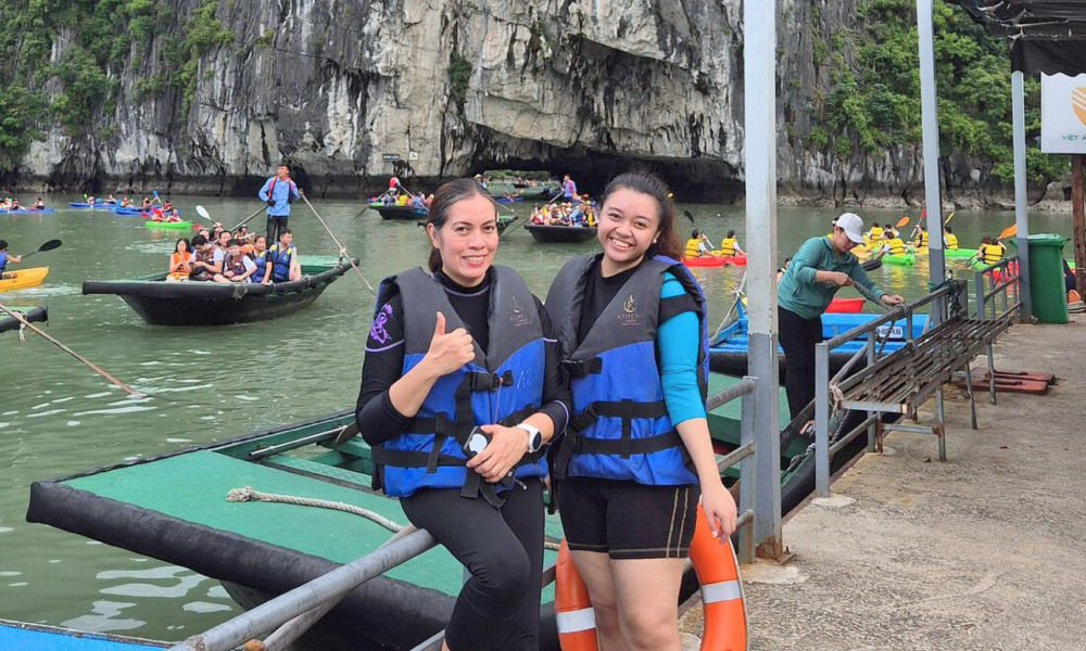 Two guests preparing to go on a bamboo boat with Halong water cave behind.