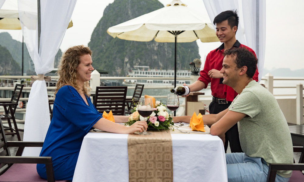 Two guests at a table on a sundeck, smiling at each other while a server pouring the wine.