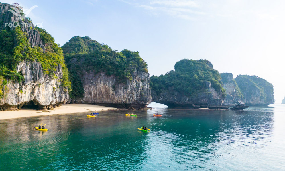 Many islands on the left with a rim of sandy shore forming a beach, with a few people kayaking to it.