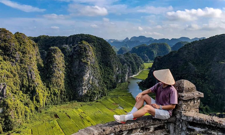 A man sitting on a fence looking out to view of Tam Coc mountains and fields.