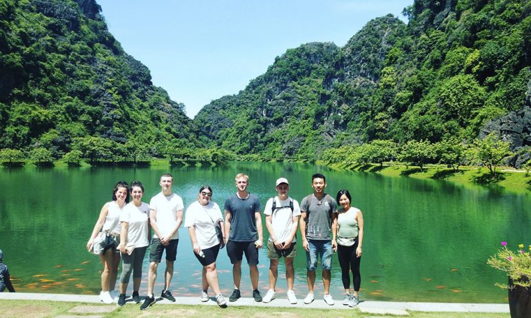 Many people posing in front of a lake and mountains behind.